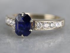 "We love the elegant style of this timeless engagement ring. The tapered, milgrain sides are interesting, and the top of the shoulders have a beautiful curve, set with bright diamonds. The center stone is a deep, excellent shade of navy blue. This color is the benchmark for fine Sapphire, and indicative of Sri Lanka, where we purchased the stone! A perfectly balanced one carat in weight and of the highest possible quality, this engagement ring will be treasured for decades to come. Metal: 14K Wh Classic Sapphire Baguette Cut Ring, Elegant Sapphire Ring With Diamond Side Stones, Timeless Sapphire Ring With Prong Setting, Timeless Sapphire Ring With Accent Stones For Formal Occasions, Classic Sapphire Birthstone Ring, Sapphire Diamond Ring With Side Stones For Promise, Classic Sapphire Halo Ring, Heirloom Style Emerald Cut Sapphire Ring With Accent Stones, Heirloom Sapphire Ring With Emerald Cut And Accent Stones