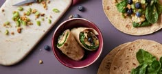 three tortillas with spinach, blueberries and other toppings on a purple surface