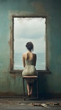 a woman is sitting on a chair looking out the window at the ocean in front of her