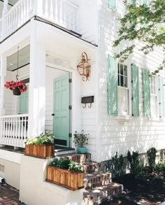 a white house with green shutters and flowers on the front porch, along with potted plants