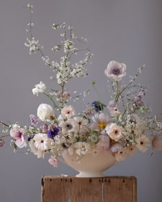 a white vase filled with lots of flowers on top of a wooden table next to a gray wall