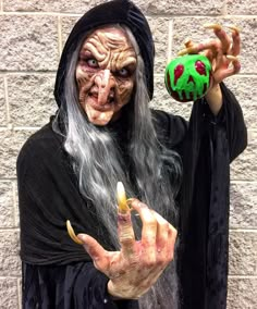 a man dressed as an old witch holding a green ball in his right hand and pointing at it