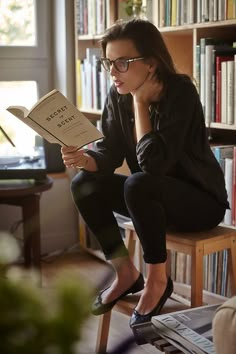 a woman sitting on a chair reading a book in front of a bookshelf