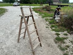 an old ladder leaning on the side of a dirt road next to a field with parked cars