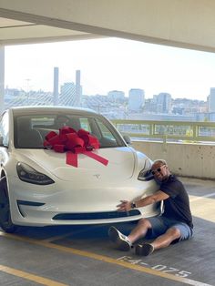 a man sitting next to a white car with a red bow on it's hood