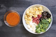 a bowl filled with pasta, broccoli and olives next to a container of tomato sauce