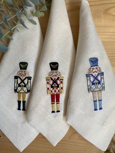 three embroidered tea towels with nutcrackers and soldiers on them, sitting next to a potted plant