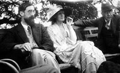 two men and a woman sitting on a park bench talking on their cell phones while the man is wearing a hat