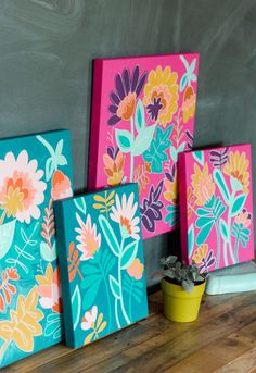 three colorful paintings on a wooden table next to a potted plant and a chalkboard