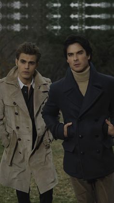 two men standing next to each other in front of a field with trees and grass