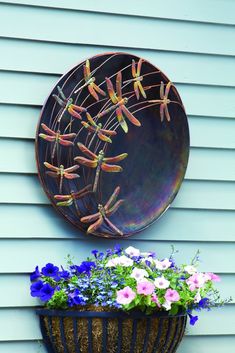 a planter filled with flowers next to a wall mounted dragonfly plate on the side of a house