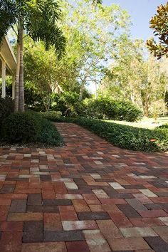 a brick walkway with trees and bushes on both sides, in front of a house