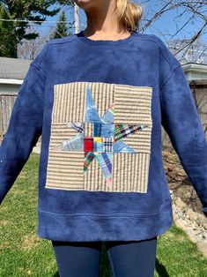 a woman standing in the grass wearing a blue sweatshirt with a patchwork star on it