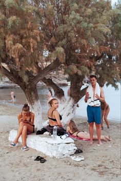 some people are sitting under a tree on the beach and one is holding a water bottle