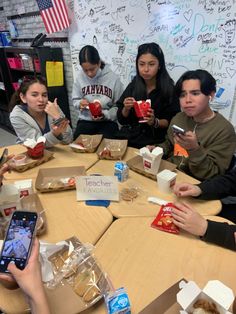 a group of young people sitting around a table eating food and drinking coffee while looking at their cell phones