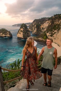 a man and woman holding hands while walking up some stairs near the ocean with cliffs in the background