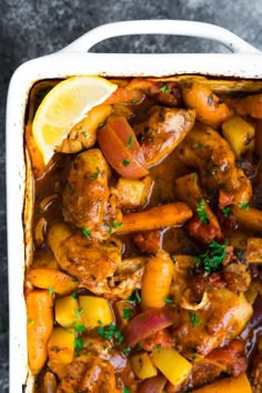 a white casserole dish filled with chicken, potatoes and carrots next to a lemon wedge