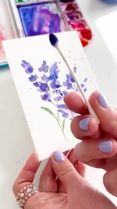 a woman holding a paintbrush in her left hand and painting on the paper with purple flowers