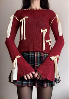 a woman wearing a red sweater with bows on her sleeves and plaid skirt is standing in front of a white wall