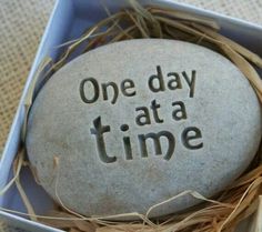 one day at a time written on a rock in a box with some dried grass