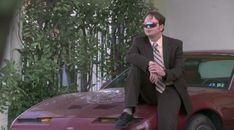 a man sitting on the hood of a red car wearing sunglasses and a suit jacket