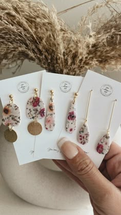 five pairs of earrings are shown in front of a white vase with dry grass and dried flowers