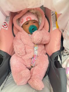 a baby in a car seat wearing a pink teddy bear outfit with pacifier attached to its mouth