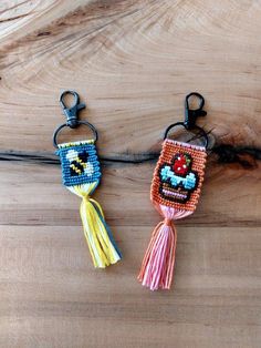 two beaded keychains are sitting on a wooden surface, one has an evil face and the other has a tassel