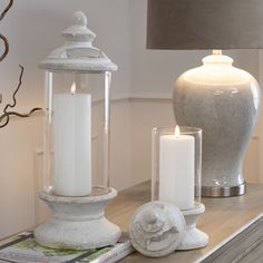 two white candles sitting on top of a wooden table next to a vase and lamp