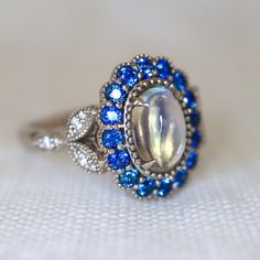 Mesmerizing Blue Moonstone Halo Ring. Moonstone engagement ring. Blue sapphires & diamonds. Edwardian Moonstone. 14K White Gold. US 7.25 A beautiful and unique moonstone ring, created with precision cut sapphires in 14K white gold, a luxurious and high-quality piece of jewelry, perfect to express your love. 💎 PREMIUM RING -- A brand new moonstone ring, personally designed and made with the highest quality of 14K white gold.  ✨ CLASSIC BRILLIANCE -- Designed with a beautiful blue moonstone at th Luxury Victorian Moonstone Ring Gift, Luxury Vintage Moonstone Ring As A Gift, Vintage Blue Sapphire Ring With Halo Setting, Vintage Blue Cluster Ring With Center Stone, Blue Vintage Cluster Ring With Center Stone, Blue Gemstone Cluster Ring Vintage Style, Vintage Blue Gemstone Cluster Ring, Antique Blue Cluster Ring, Vintage Sapphire Cluster Ring With Gemstones