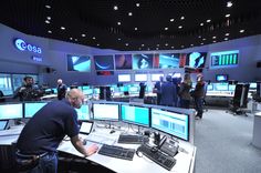 a man sitting at a desk in front of multiple computer monitors with people standing around