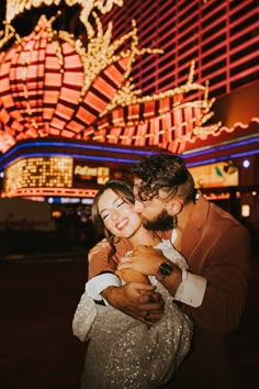 a man and woman hugging in front of a building at night with neon lights behind them