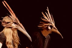 two people with long hair wearing masks made out of sticks and feathers on their heads