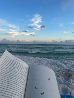 an open book sitting on top of a sandy beach next to the ocean under a blue cloudy sky