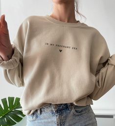 a woman wearing a beige sweatshirt with the words in my pregnant era printed on it