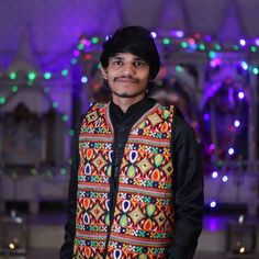 a man standing in front of a stage wearing a colorful vest and black shirt with lights behind him