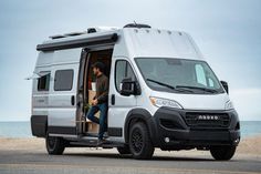 a man standing in the open door of a white van near the ocean and sand