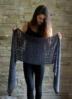 a woman holding up a crocheted shawl in front of a brick wall