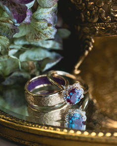 A matching wedding ring set from Staghead Designs sits on a mirror surrounded by flowers and gold trinkets. This ring set features our "Jane" floral engagement ring in 14K yellow gold with an oval lab-grown alexandrite center stone and diamond accents, paired with a 6mm width men's wedding band in hammered 14K yellow gold with sleepy lavender opal lining. Pearl And Alexandrite Ring, Dark Opal Engagement Ring, Vintage Alexandrite Engagement Rings, Purple Rings Engagement, Lavender Wedding Ring, Hammered Gold Wedding Band, Alexandrite Engagement Rings, Opal Wedding Set, Opal Wedding Ring Set