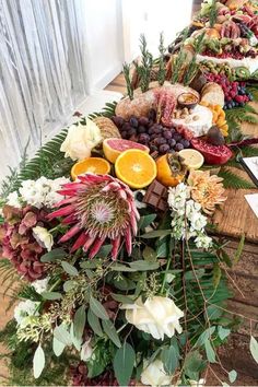 an arrangement of fruits and flowers on a long wooden table in front of a window