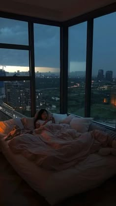 a woman is laying on her bed in front of large windows with city lights at night