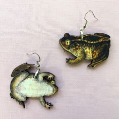 two frog shaped earrings hanging from hooks on a white surface, one is green and the other is brown