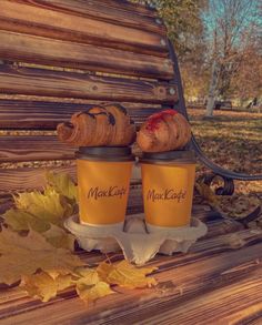 two coffee mugs sitting on top of a wooden bench next to leaves and fallen leaves