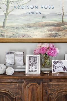 pink flowers are in a vase next to pictures on a wooden cabinet with a painting behind it