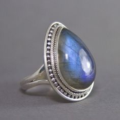 SKU: SS-007Material: 925 stamped sterling silverGemstone(s): Labradorite (N)Gemstone Dimensions: 15 X 22 X 5 mmRing Size: US 7.5Weight: 9.4 gramsThis breathtaking Labradorite cabochon ring is truly a beauty! Set in a classic bezel with a granulated border, the setting enhances the large Labradorite beautifully. The Labradorite not only has a strong blue fire that can be seen from every angle, it also has mica flakes, which add that extra shimmer to an already incredible stone! Silver Teardrop Cabochon Rings, Teardrop Moonstone Ring In Sterling Silver, Silver Teardrop Gemstones For Anniversary, Silver Pear-shaped Moonstone Ring, Sterling Silver Teardrop Moonstone Ring, Silver Teardrop Moonstone Ring In Sterling Silver, Silver Teardrop Ring With Stone Setting, Silver Teardrop Gemstones, Silver Teardrop Rings With Spiritual Style