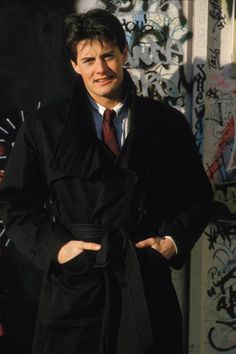 a man in a coat and tie standing next to a wall with graffiti on it