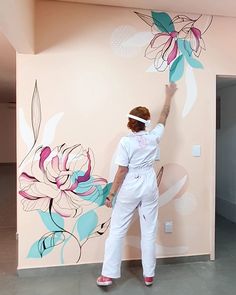 a woman in white jumpsuit painting a wall with flower designs on it and pink walls behind her