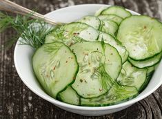 a white bowl filled with sliced cucumbers and herbs next to a wooden table