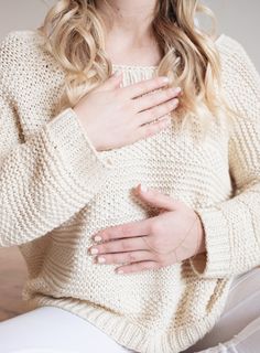 a woman with her hands on her chest wearing a white sweater and jeans, sitting down