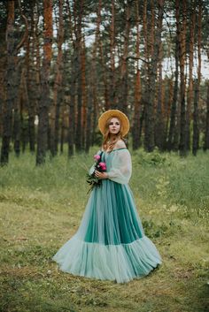 a woman in a long dress and straw hat standing in the woods with her hands on her hips